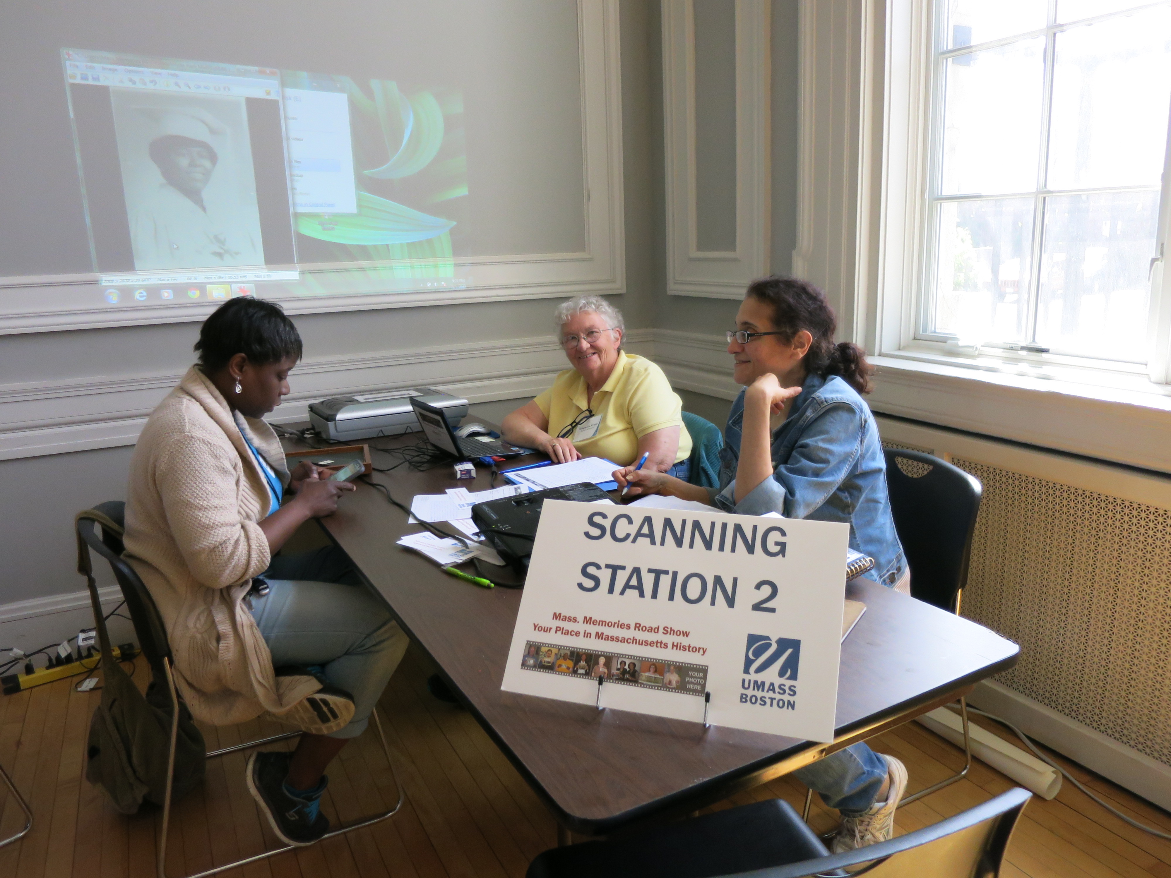 <p>Scanning Station volunteers assist a participant with her photo at the Hyde Park (Boston) Mass. Memories Road Show, 2016. Thoughout RoPA, the Scanning Station is called the Copying Station.</p>