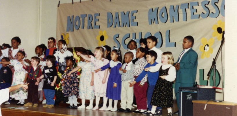 A number of students in a line on stage in front of a banner that reads Notre Dame Montessori School